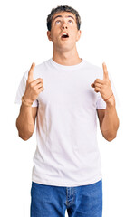 Naklejka premium Young handsome man wearing casual white tshirt amazed and surprised looking up and pointing with fingers and raised arms.