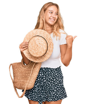 Young blonde girl holding summer hat and wicker handbag pointing thumb up to the side smiling happy with open mouth