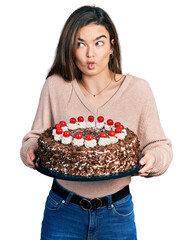 Young caucasian girl celebrating birthday holding big chocolate cake making fish face with mouth and squinting eyes, crazy and comical.