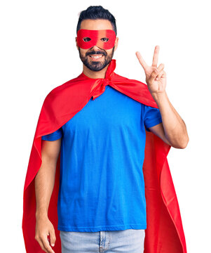 Young hispanic man wearing super hero costume smiling with happy face winking at the camera doing victory sign. number two.