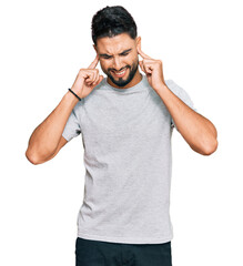 Fototapeta premium Young man with beard wearing casual grey tshirt covering ears with fingers with annoyed expression for the noise of loud music. deaf concept.