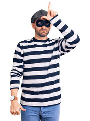 Young handsome man wearing burglar mask making fun of people with fingers on forehead doing loser gesture mocking and insulting.