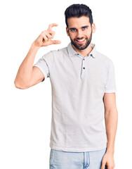 Young handsome man with beard wearing casual polo smiling and confident gesturing with hand doing small size sign with fingers looking and the camera. measure concept.