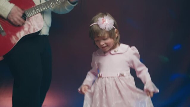 Little girl on stage in pink vintage dress, she dances, her father plays acoustic guitar. Color music is shining. Performance on stage. Early development of children