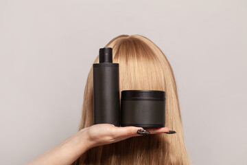 Plastic bottles of hair care and beauty products in female hand against blonde head.