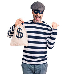 Middle age handsome man wearing burglar mask holding money bag pointing thumb up to the side smiling happy with open mouth