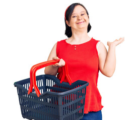 Brunette woman with down syndrome holding supermarket shopping basket celebrating victory with happy smile and winner expression with raised hands