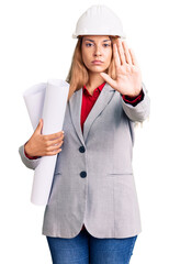 Beautiful young woman wearing architect hardhat holding build project with open hand doing stop sign with serious and confident expression, defense gesture