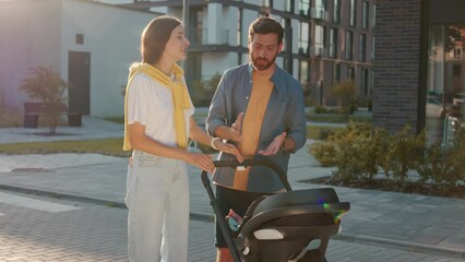 Footage of beautiful young couple in casual clothes standing near building with baby stroller in modern residential complex. Husband and wife quarreling, gesturing. Father taking baby out of baby