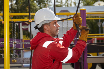 A specialist repairs the playground. A specialist in a white construction helmet and a red jumpsuit.