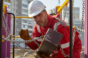A specialist repairs the playground. A specialist in a white construction helmet and a red jumpsuit.	