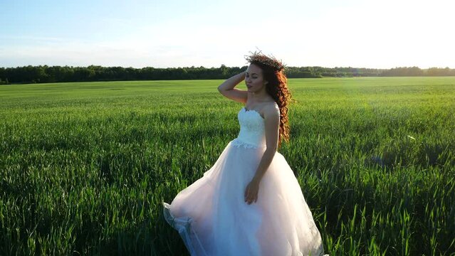 Bride fixes her wreath on hair while standing in the middle of wheat field. Wedding dress sways in the wind.