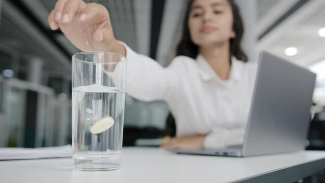 Young overworked manager woman suffering from headache feels migraine symptom feeling unwell from fatigue unhealthy girl sitting in office throwing soluble painkiller pill into water focused on remedy