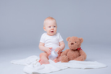 a baby boy in a white bodysuit sits with a teddy bear on a white background, holding a bottle of...