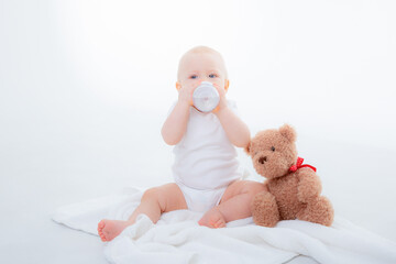 a baby boy in a white bodysuit sits with a teddy bear on a white background, holding a bottle of water , drinking water