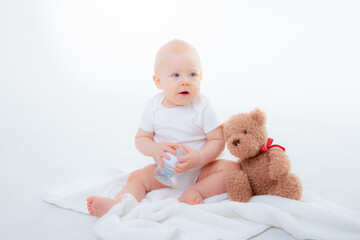 a baby boy in a white bodysuit sits with a teddy bear on a white background, holding a bottle of water , drinking water