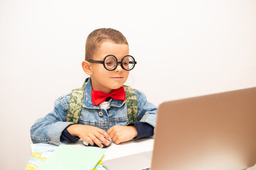 Cute little schoolboy in nerd glasses with a red bow tie sits at a table with a laptop