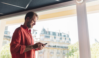 Caucasian man (businessman) using a phone (smartphone) next to the big office window - corporation.