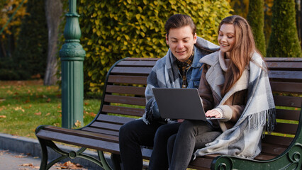 Fototapeta premium Couple freelancers two partners people young caucasian woman and man entrepreneurs sit on bench in park use urban wifi work on laptop look at device screen make internet order discuss business startup