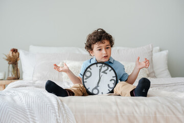 Curly handsome Spanish boy sitting on bed with big wall clock spreading hands in disbelief expression, waiting parents to spend time together. Kids psychology concept. Mockup kids growing time.