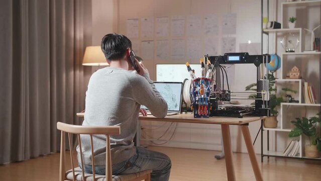 Back View Of A Male With 3D Printing Talking On Smartphone While Designing A Cyborg Hand On A Laptop At Home
