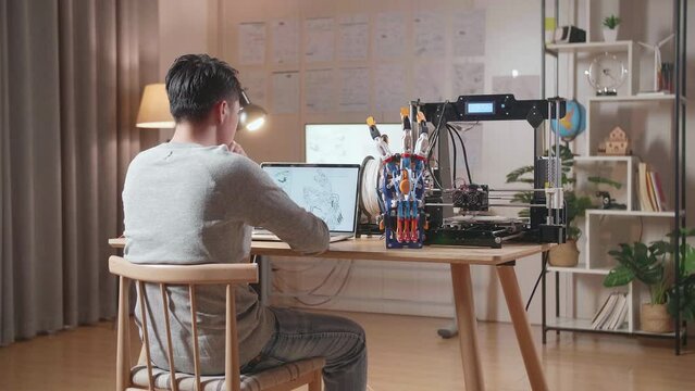 Back View Of A Male With 3D Printing Thinking While Designing A Cyborg Hand On A Laptop At Home
