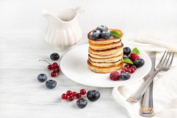 Appetizing pancakes with berries and powdered sugar on a light plate, a fork and a knife on a light table.