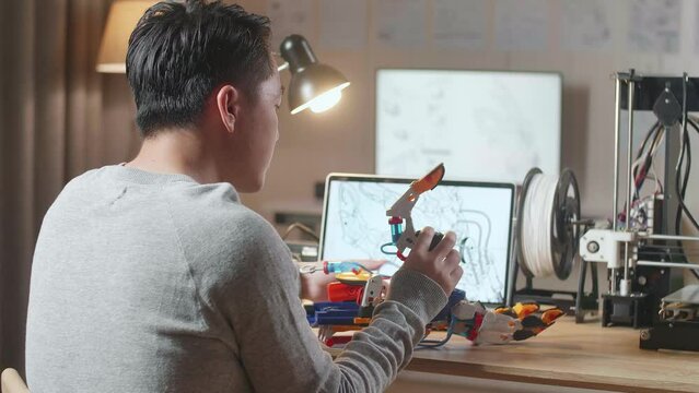 Side View Of Asian Male With 3D Printing Assembling A Cyborg Hand While Designing It On A Laptop At Home
