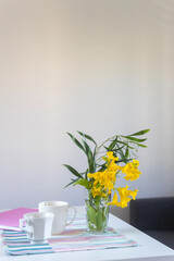 Shrub branch with Tecoma stans flowers in a glass beaker on a white table in the interior. A pink spring notepad, two coffee mugs and a geometric patterned napkin.