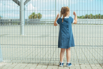 a little preschooler girl in a blue dress stands near a barrier from a high mesh fence and holds on...