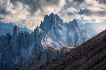 Obraz premium Epic rocky mountains at sunset in autumn. Mountain landscape with alpine mountains, orange grass on the hill, dramatic cloudy sky in fall. Aerial view of high rocks. Tre Cimein Dolomites, Italy