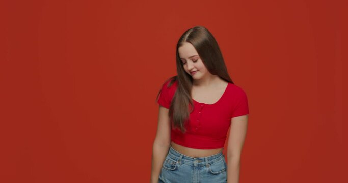 Embarrassed shy young girl smiling posing flirting fixes her hair, makes eyes on orange studio background. Copy space