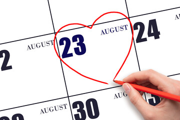 A woman's hand drawing a red heart shape on the calendar date of 23 August. Heart as a symbol of...