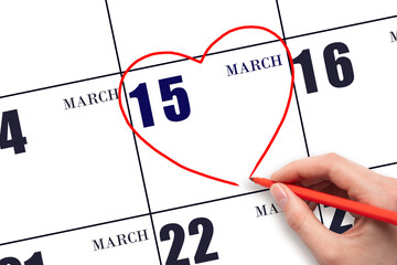 A woman's hand drawing a red heart shape on the calendar date of 15 March. Heart as a symbol of...
