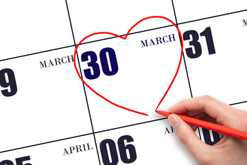 A woman's hand drawing a red heart shape on the calendar date of 30 March. Heart as a symbol of...