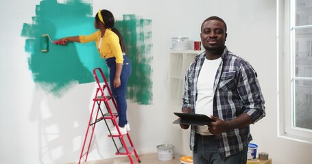 Portrait of handsome young African American man standing in room typing on tablet device browsing online while wife painting wall in green color. Apartment improvement, repair, renovation concept