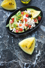 Yellow watermelon, goat cheese and tomato salad, above view on a black marble background, vertical shot with space