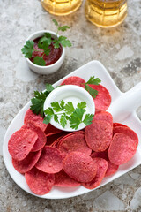 Shovel-shaped plate with red potato chips, beer and dips, vertical shot on a light-brown granite background, high angle view
