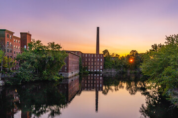 A complex of historic cotton mill buildings with a chimney in an old industrial park with a reflection in the Nashua River late at sunset. Nashua, New Hampshire, USA