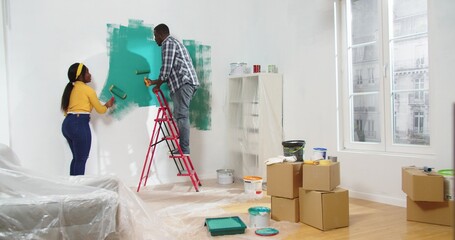 Rear of African American young married couple wife and husband painting wall in green color using...