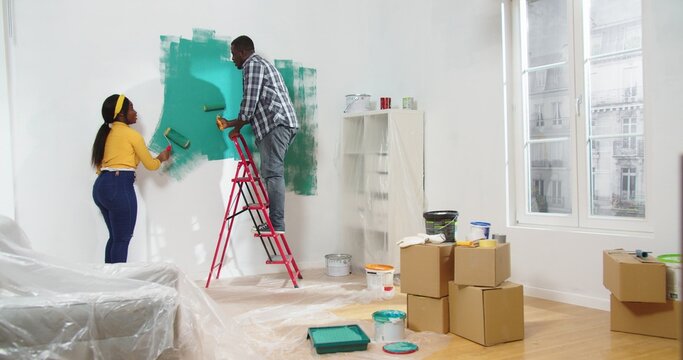 Rear of African American young married couple wife and husband painting wall in green color using paint roller brush in new house with boxes. Apartment renovation, improvement, decoration concept