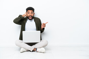 Young Brazilian man with a laptop sitting on the floor isolated on white surprised and pointing finger to the side