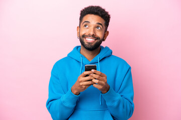 Young Brazilian man isolated on pink background using mobile phone and looking up