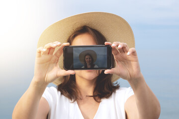 A young girl in a white dress and a straw hat stands against the background of the sea takes a selfie.