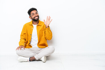 Young Brazilian man sitting on the floor isolated on white background saluting with hand with happy expression © luismolinero