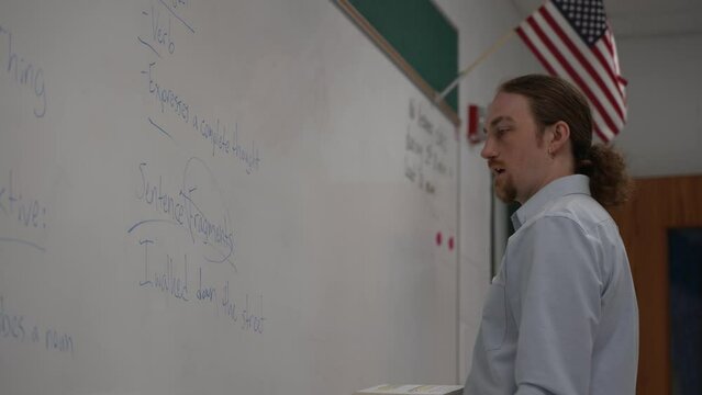 Male school teacher holding book teaching english in school classroom at a whiteboard, with US flag.