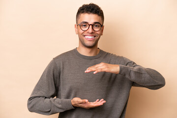 Young Brazilian man isolated on beige background holding copyspace imaginary on the palm to insert...