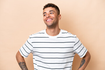 Young Brazilian man isolated on beige background posing with arms at hip and smiling