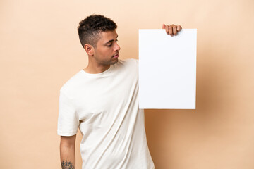 Young Brazilian man isolated on beige background holding an empty placard and looking it