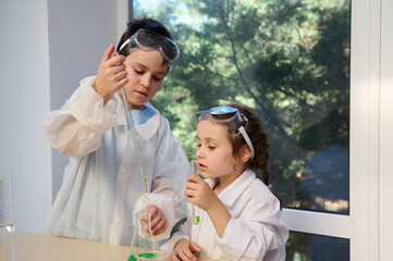 Adorable elementary children, boy and girl in white lab coats and safety goggles, making science experiments in chemistry class. Back to school on the new semester of the academic year. New knowledges